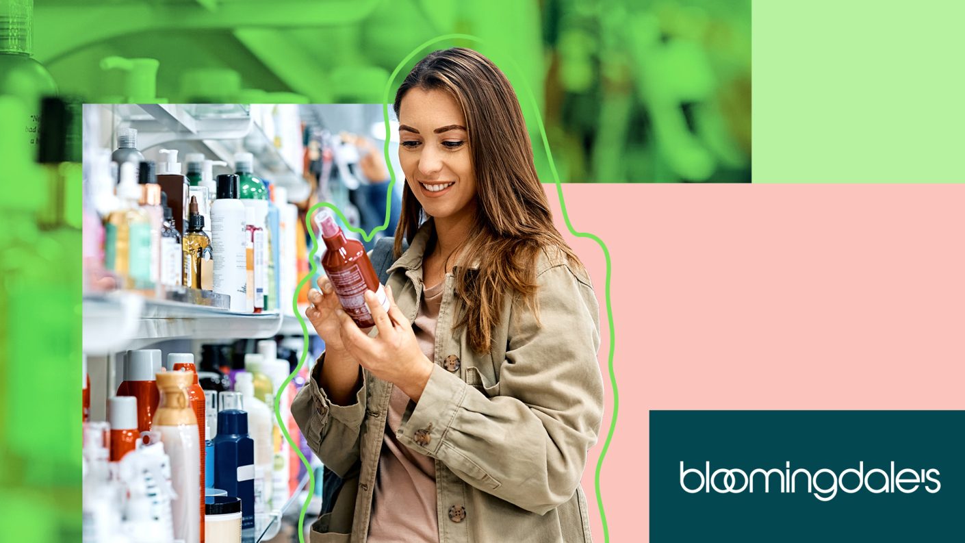 Woman with brown hair holding a spray bottle in her hand, looking at the beautyproduct, in front of a shelf filled with beauty products